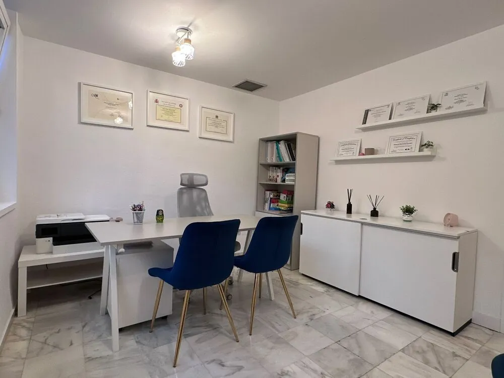 Despacho con mesa blanca, dos sillas azules y títulos enmarcados en la pared